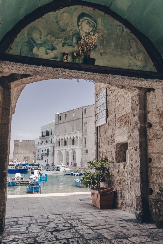Porte d'entrée du port de Monopoli avec la fresque peinte d'une Vierge, des bateaux colorés, de l’eau calme et des bâtiments en pierre claire sous un ciel dégagé.